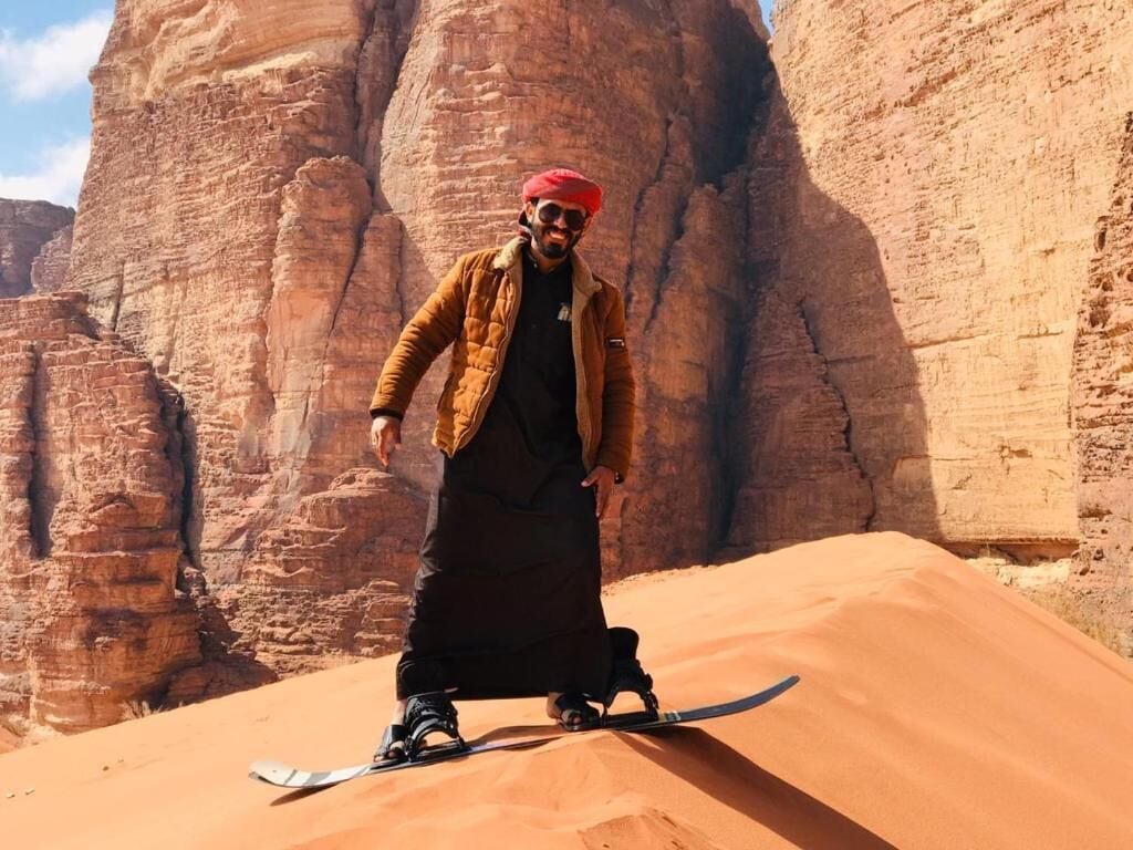 Uma pessoa em cima de uma prancha de snowboard em um deserto, com formações rochosas ao fundo.