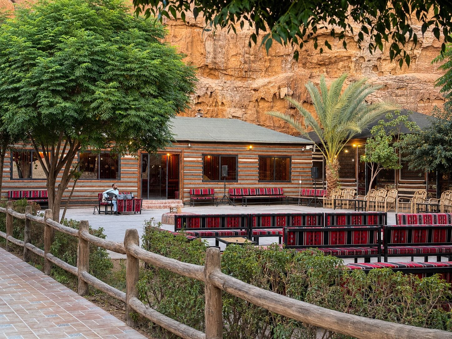 A rustic building with a wooden exterior, outdoor seating with red cushions, and a stone wall in the background.