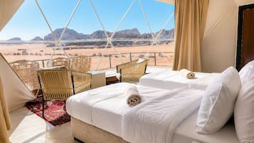 A tented room with a bed, two chairs, a small table, and a view of a desert landscape.