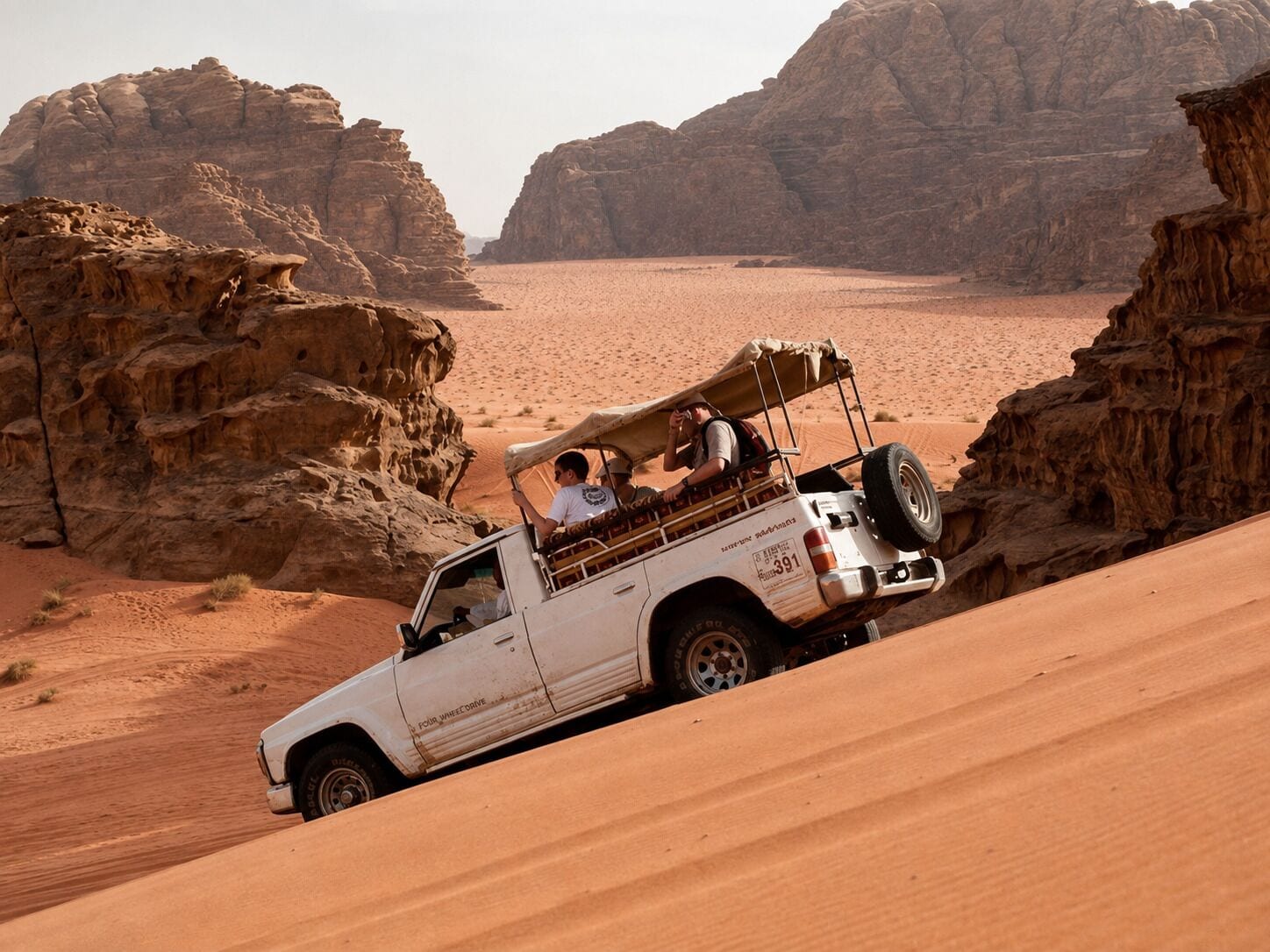 A white off-road vehicle with two passengers, navigating a sandy terrain between rocky cliffs.