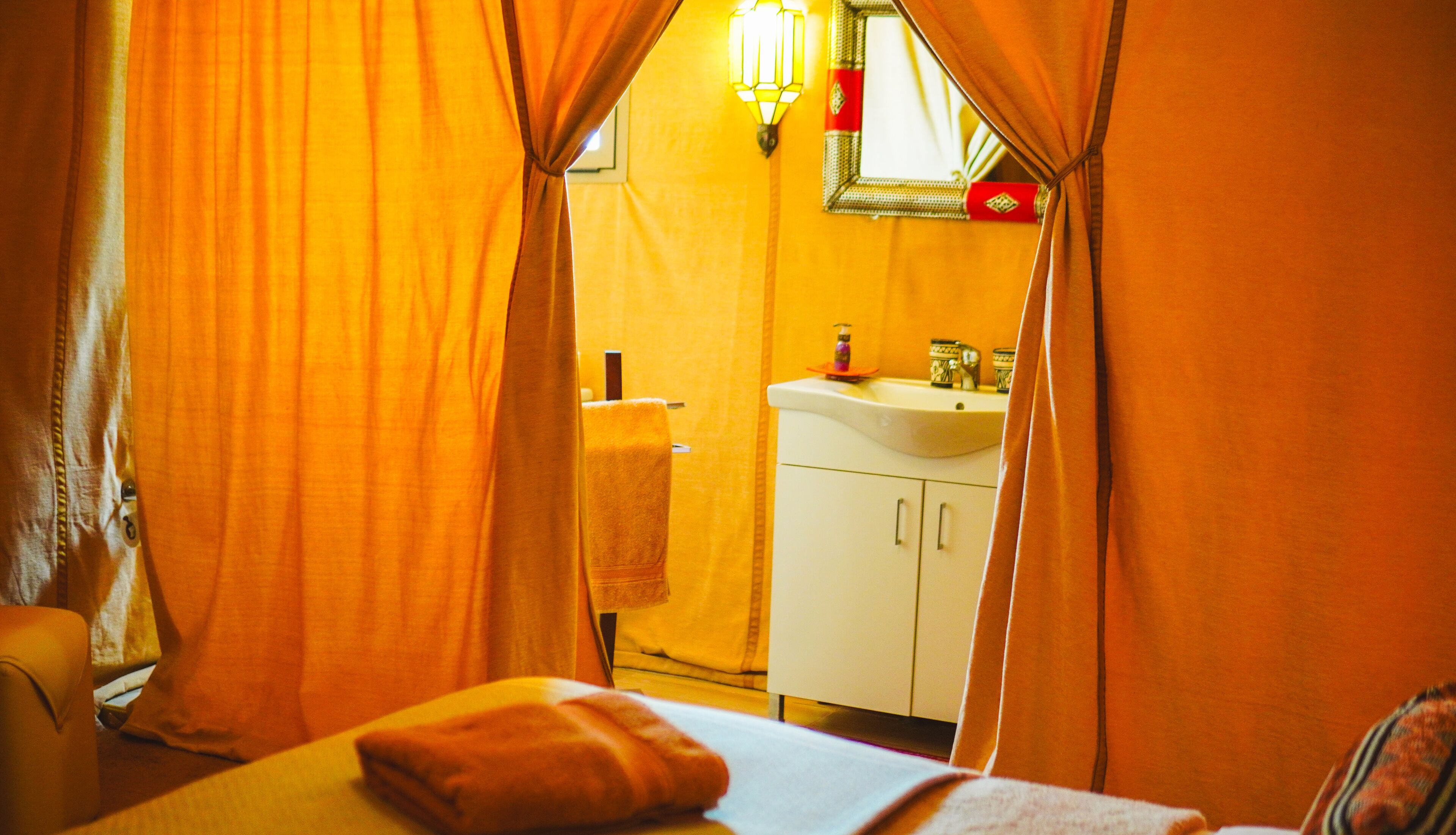 A room with orange curtains, a bed with a pillow, a white sink, and a mirror above it.