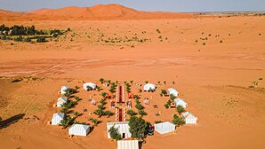 An aerial view of a desert camp with several white tents arranged in a circular pattern, surrounded by sparse vegetation and distant sand dunes. - Saharian Luxury Camp (Rissani)