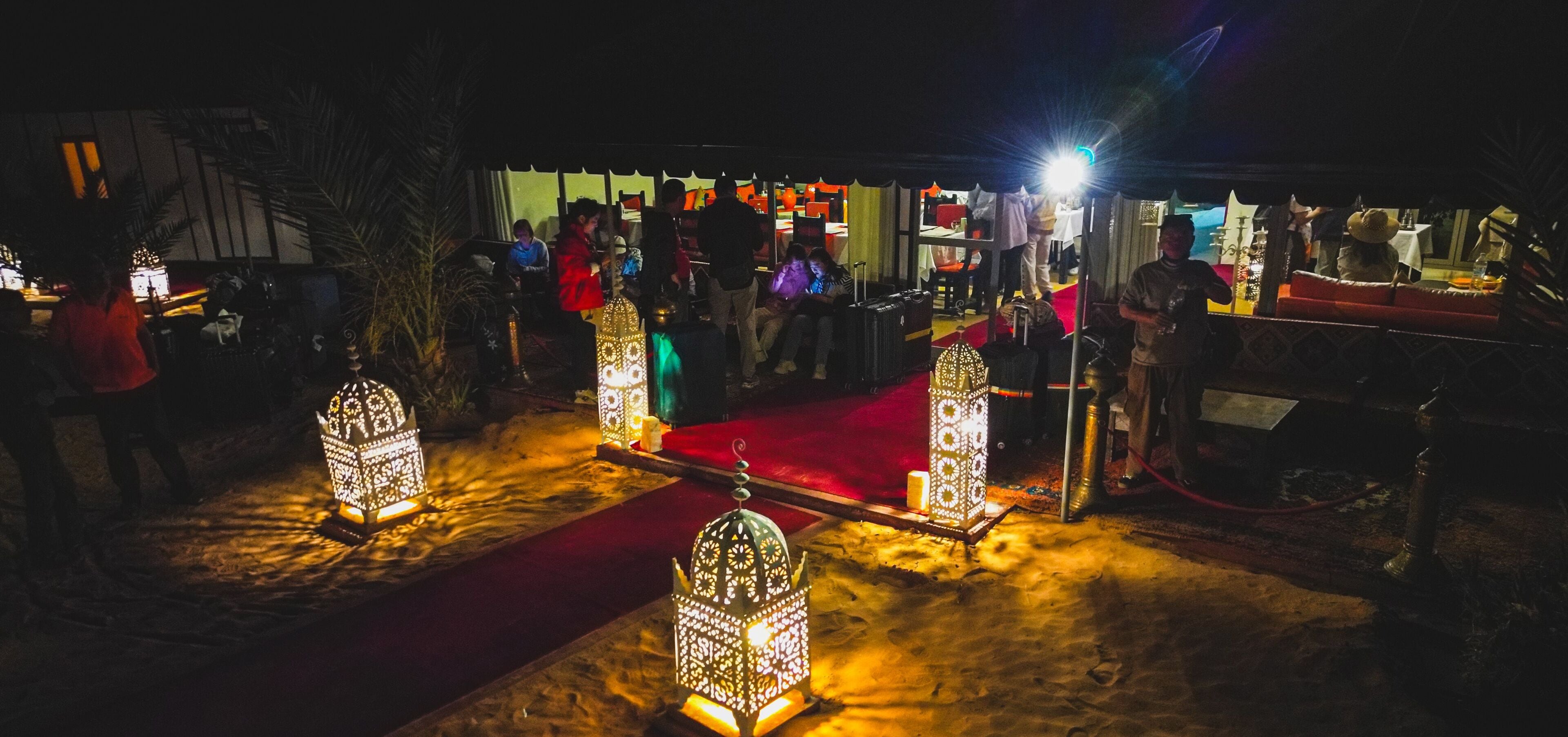 A nighttime outdoor area with ornate lanterns, a red carpet, and people gathered around tables and chairs.
