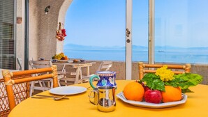 A table set with fruits, a mug, and a plate, overlooking a body of water with a distant ship.