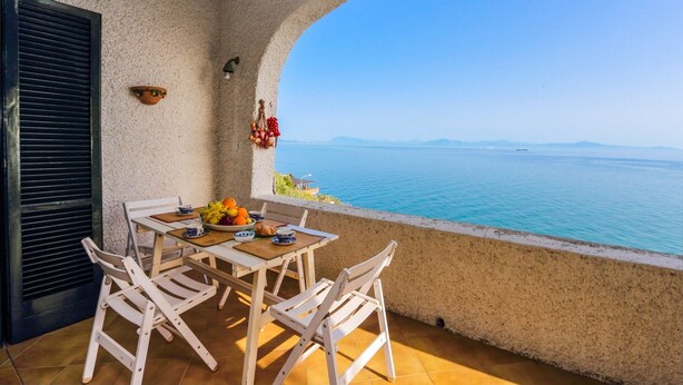 A balcony with a table set for two, overlooking a calm sea and distant mountains.