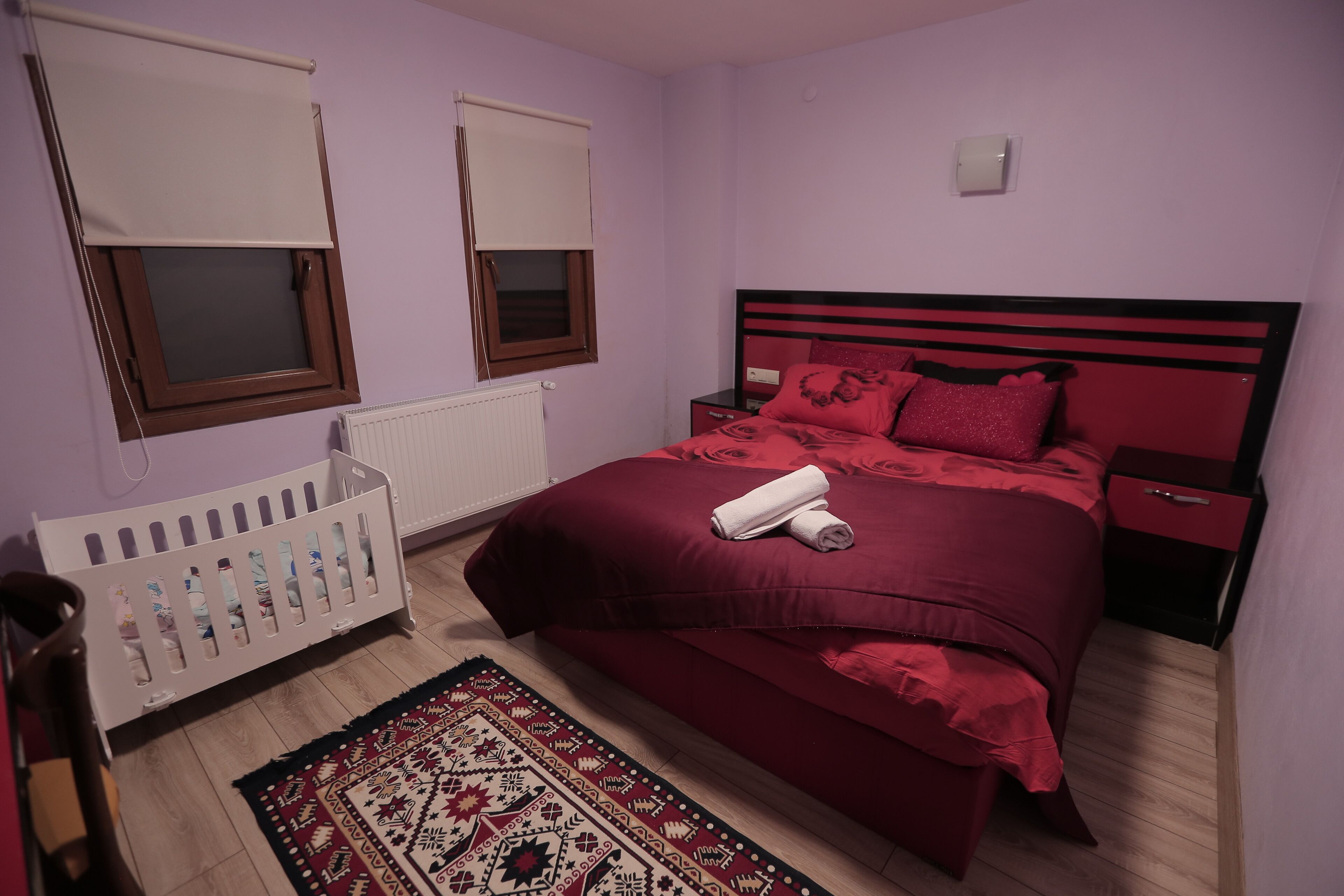 Un dormitorio con una cama roja, una cuna blanca y una alfombra estampada.