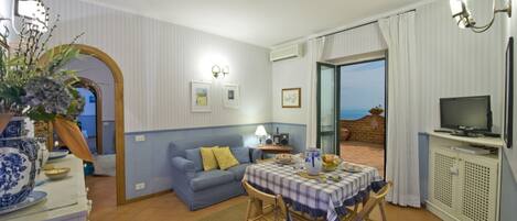 Una sala de estar con un sofá, una mesa de comedor con mantel a cuadros, un televisor y vista al mar a través de un ventanal.