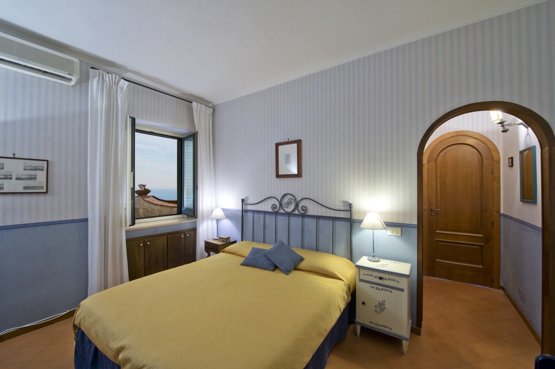 Ein Hotelzimmer mit einem Bett, einem Nachttisch, einer Lampe, einem Fenster mit Vorhängen, einer Tür und einem gerahmten Bild an der Wand.