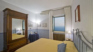 Quarto de hotel com uma cama grande, um guarda-roupa de madeira, uma escrivaninha com abajur, uma janela com vista e quadros nas paredes.