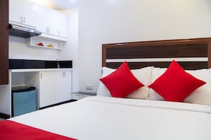 Habitació d'hotel amb llit, parets blanques, capçalera de fusta i una zona de cuina americana amb armaris i fogó.