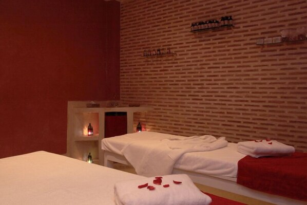 Une salle de spa avec deux lits, chacun agrémenté d’une serviette blanche et de pétales de rose, et un mur de briques orné d’étagères remplies de bouteilles.