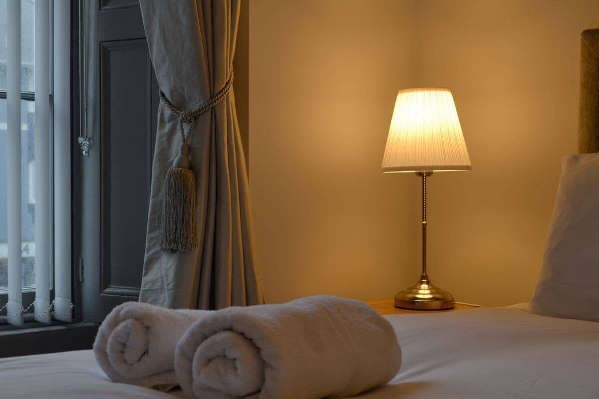 Una cama bien hecha con toallas enrolladas, una lámpara de noche y una ventana con cortinas.
