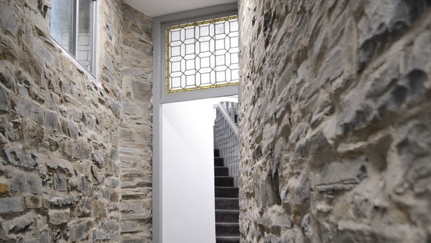 Ein schmaler Stein-Korridor mit einer Treppe, die zu einem Raum mit weißer Wand und einem Buntglasfenster führt.