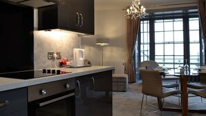 Eine moderne Küche mit dunklen Schränken, einem eingebauten Backofen und einer Arbeitsplatte mit Wasserkocher und Kaffeemaschine. Es gibt eine Essecke mit Tisch und Stühlen sowie ein großes Fenster mit Vorhängen.