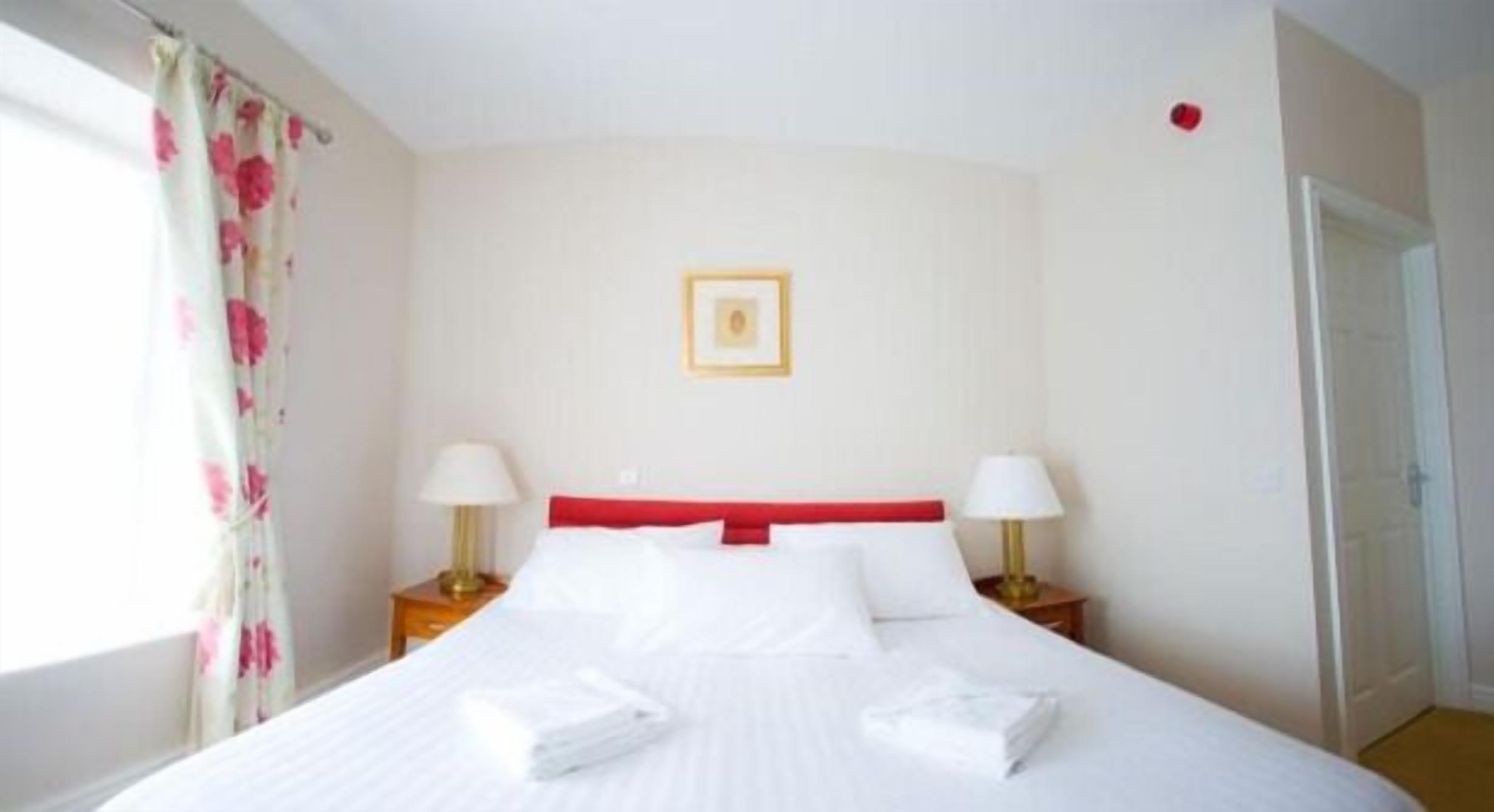 Habitación de hotel con cama, dos mesitas de noche con lámparas, cabecero rojo, un cuadro en la pared y una ventana con cortinas.
