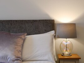 Una cama con cabecera gris, almohada blanca y lámpara sobre una mesita de noche de madera.