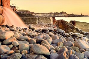 A rocky shoreline with a waterfall, a stone wall, and a distant cityscape. - Queensbridge Hotel (Aberystwyth)