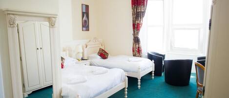 Une chambre avec deux lits blancs, une grande fenêtre avec des rideaux, une armoire et une chaise.
