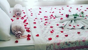 A neatly made bed with rose petals and a bouquet of flowers, a remote control, and a decorative glass on a bedside table. - Hidirlik Pansiyon (Antalya)