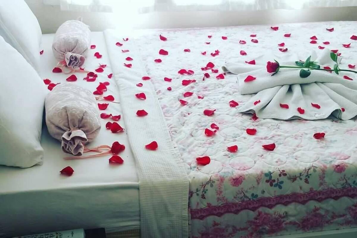 A neatly made bed with rose petals and a bouquet of flowers, a remote control, and a decorative glass on a bedside table.