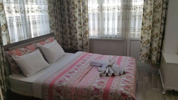 A neatly made bed with a floral bedspread and pillows, a bedside table, and curtains with a floral pattern. - Hidirlik Pansiyon (Antalya)