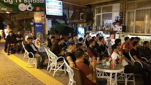 A crowded outdoor seating area with white plastic chairs and tables, a TV screen, and a bar with bottles. - Hidirlik Pansiyon (Antalya)