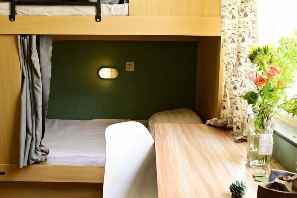 Una cama litera con una pared verde, una mesita de noche, una lámpara pequeña y una ventana con cortinas.