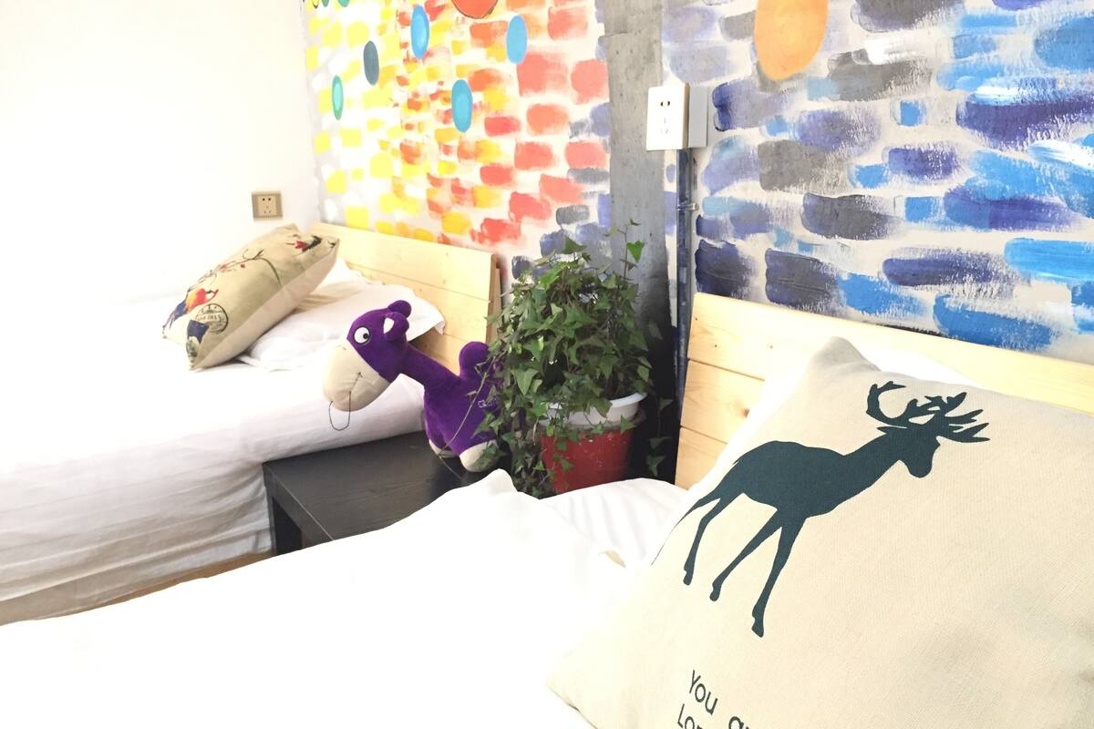Un dormitorio con dos camas, una pared con arte abstracto de colores, una planta en maceta y una almohada con un diseño de ciervo.