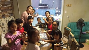 A group of people sitting around a wooden dining table in a room with a kitchen in the background. - Weihai Hiking International Youth Hostel (Weihai)