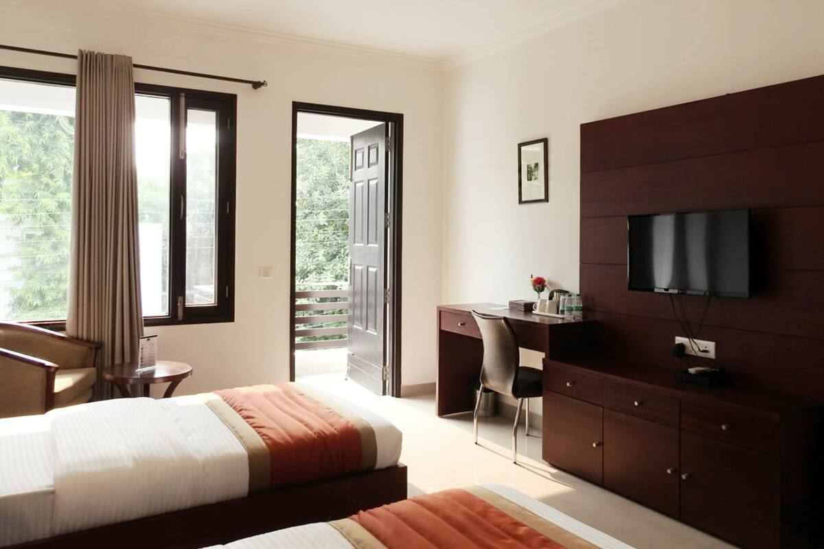 Habitación de hotel con cama, escritorio, silla, televisor y balcón con cortinas.