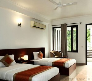 A hotel room with two beds, a ceiling fan, an air conditioning unit, a nightstand with a lamp, a chair, and a view of greenery outside. - Hotel Spoontree Boutique BnB (Gurugram)