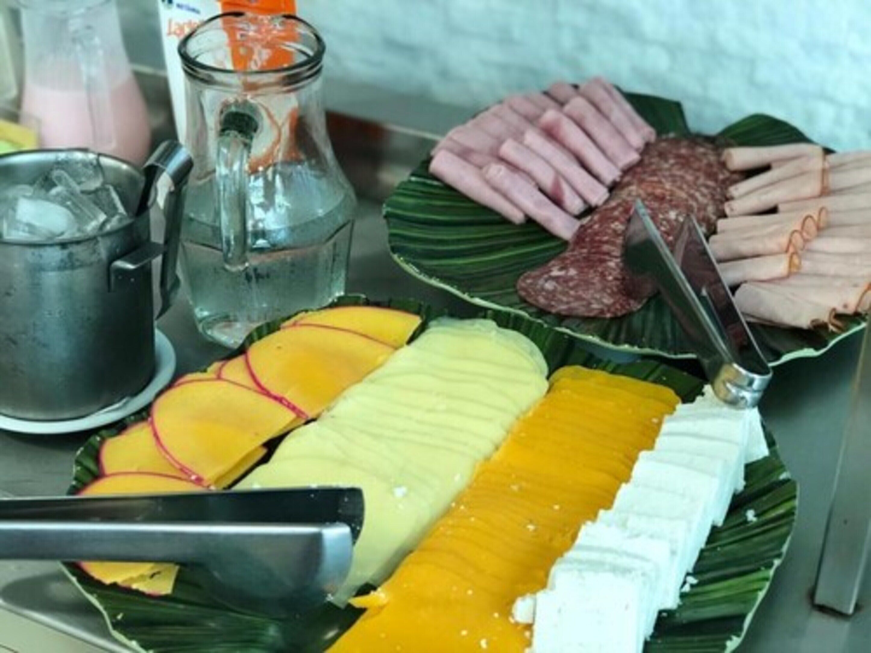 Variedad de alimentos en un plato, incluyendo mango en rodajas, queso blanco y embutidos, con un cuchillo y una jarrón de vidrio al fondo.