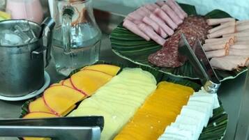 Variedad de alimentos en un plato, incluyendo mango en rodajas, queso blanco y embutidos, con un cuchillo y una jarrón de vidrio al fondo.