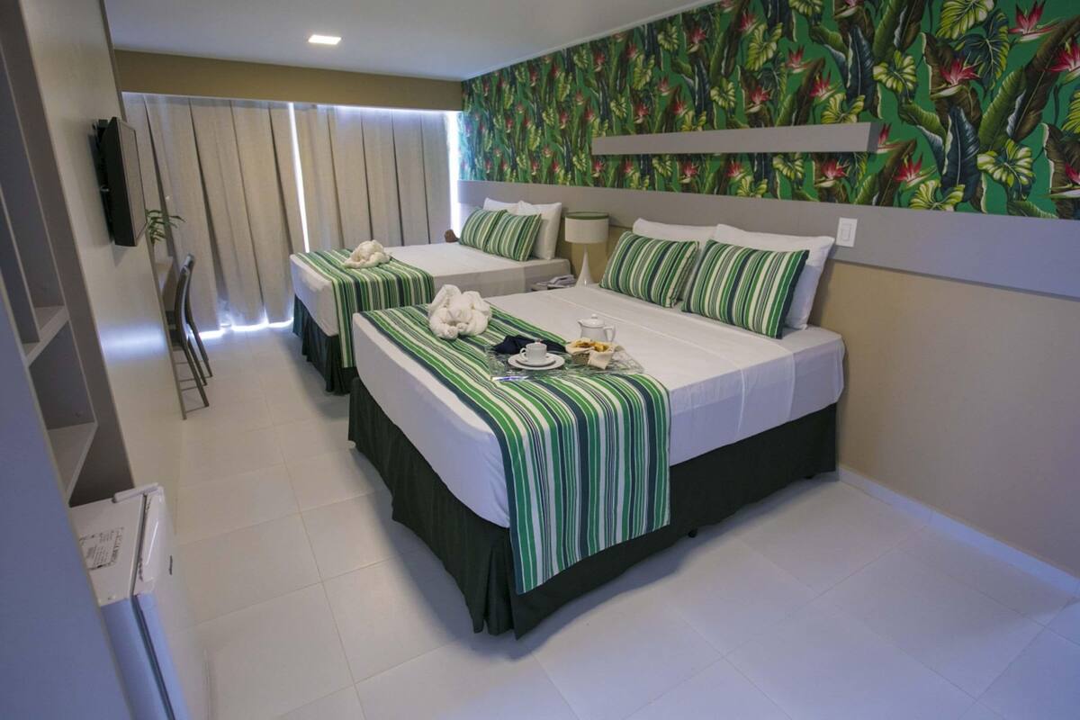 A hotel room with two beds, a television, a window with curtains, and a tropical wallpaper.