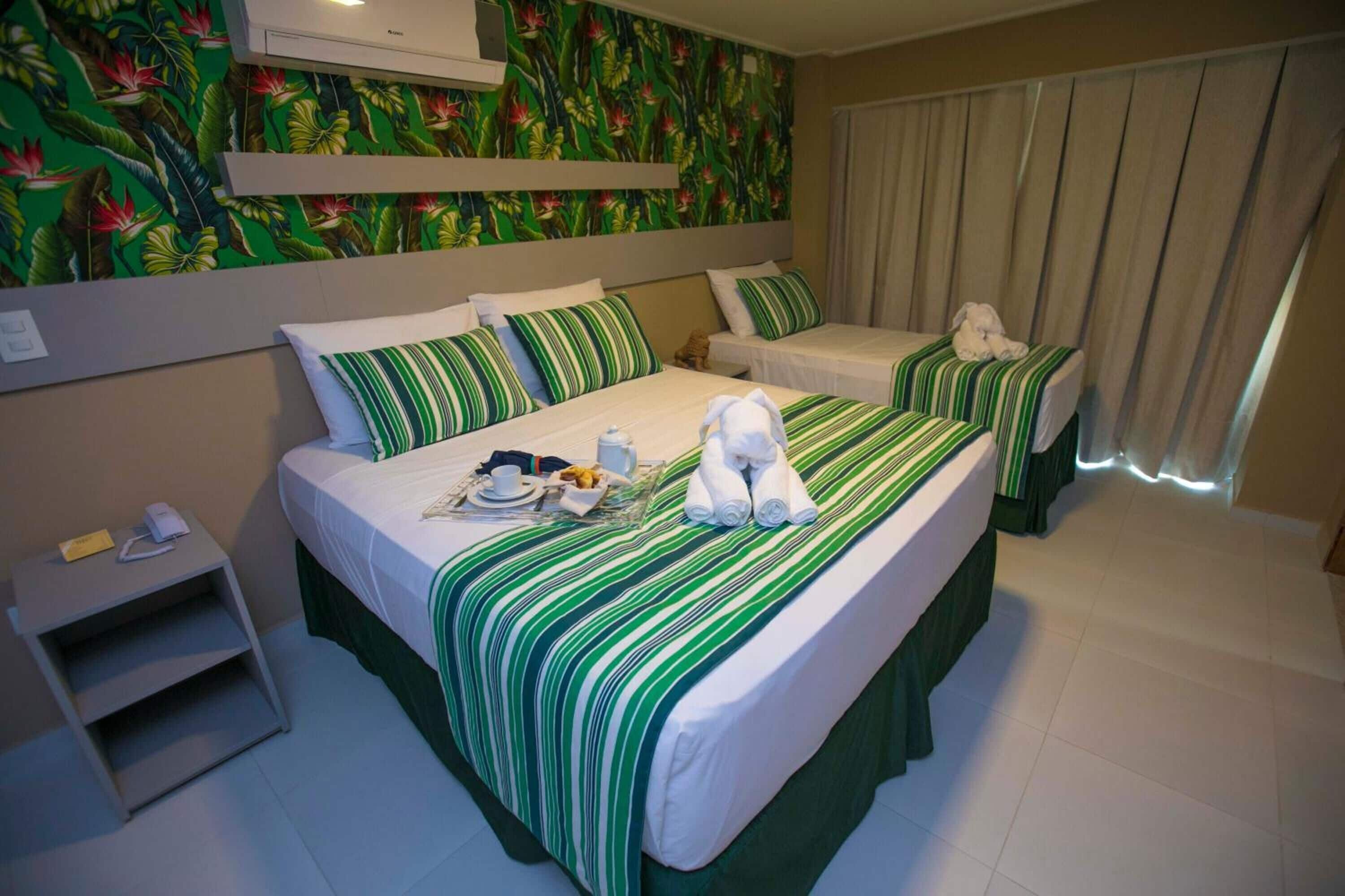 A hotel room with two beds, each with green-striped bedding, a small bedside table, and a wall with tropical wallpaper.