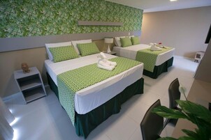 Habitación de hotel con dos camas, cada una con ropa de cama verde estampada y almohadas blancas. Hay una mesita de noche pequeña con una lámpara, una silla y una planta.