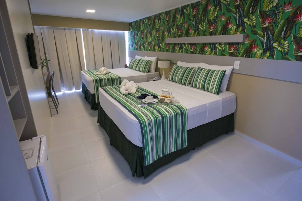 A hotel room with two beds, a TV, a window with curtains, and a tropical wallpaper.