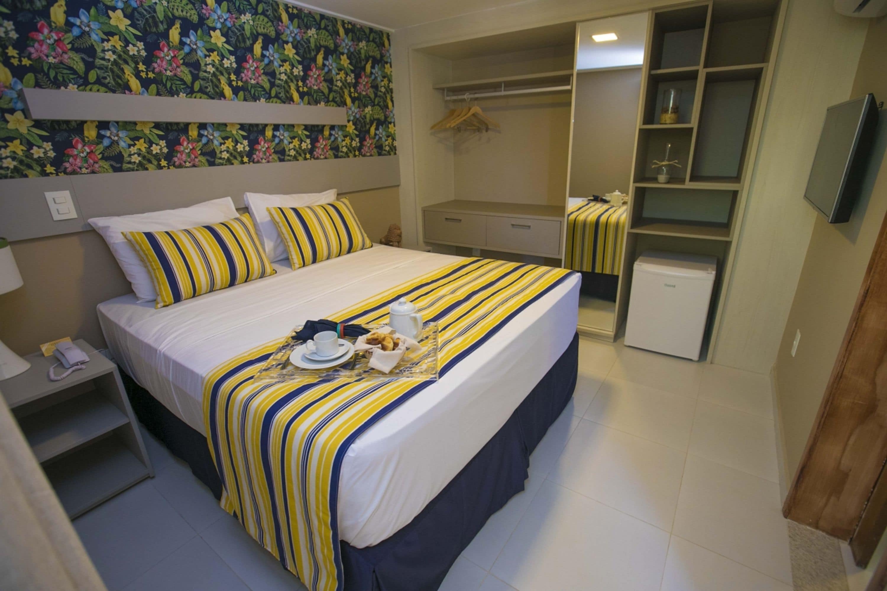 A neatly made bed with striped bedding, a bedside table with a phone and a cup, and a small refrigerator.