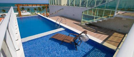 A rooftop pool with a glass balustrade, a lounge chair, and a view of the ocean.