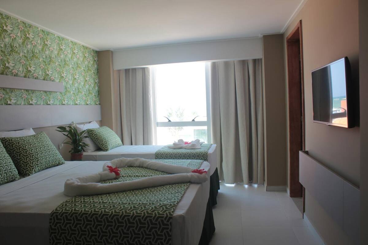 A hotel room with two beds, a TV, a potted plant, and a view of the outdoors.