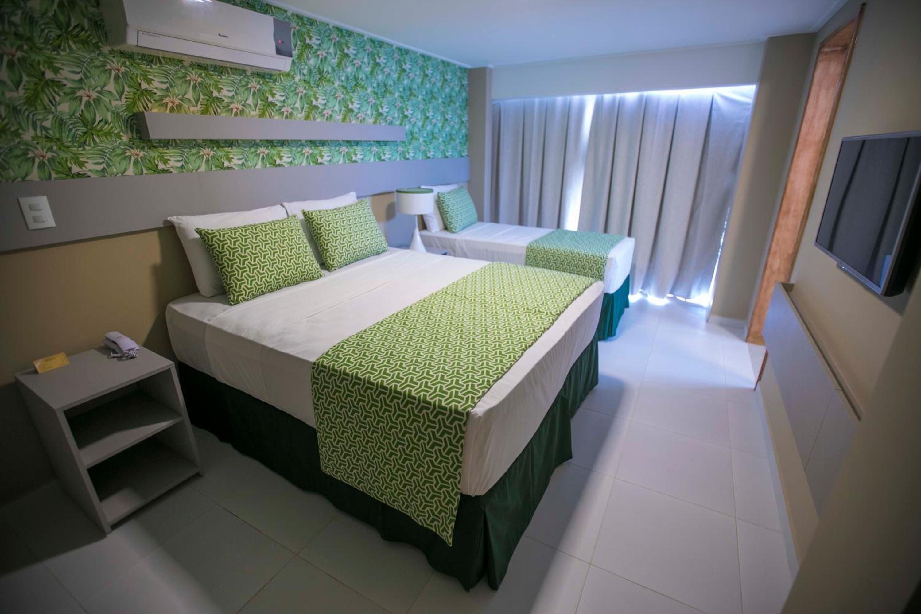 A hotel room with a large bed, two smaller beds, a TV, a desk, and a green patterned wall.