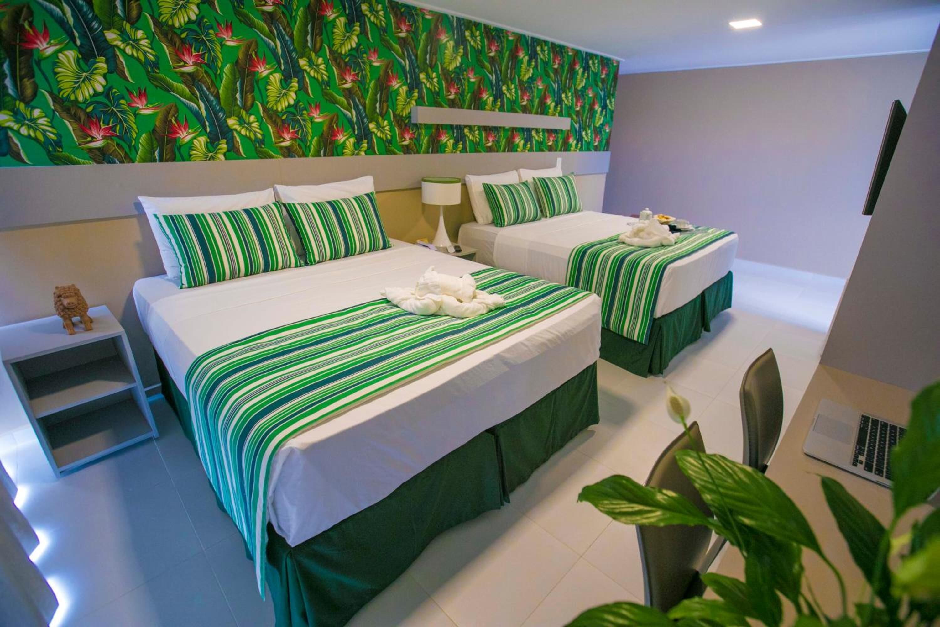 A hotel room with two beds, each with green-striped bedding and white pillows. There is a nightstand with a lamp, a small table with a teddy bear, and a laptop on the floor.