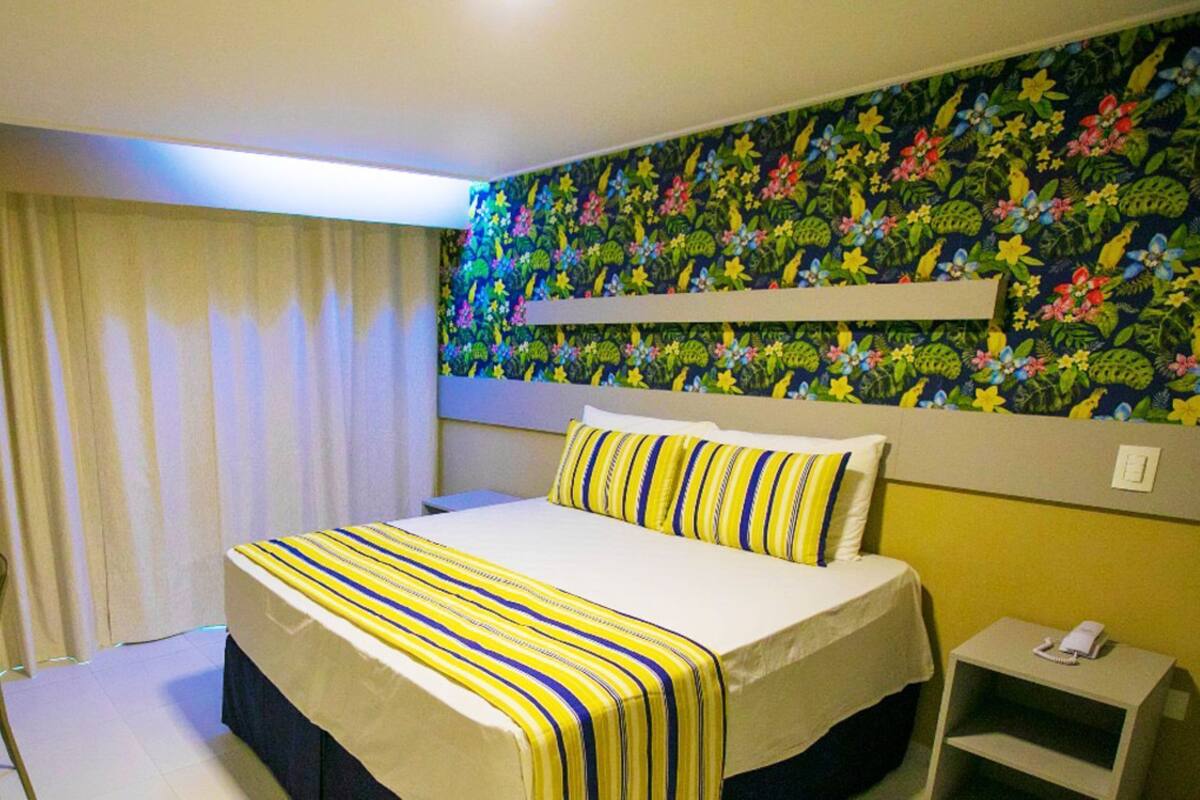 A hotel room with a bed, a desk with a laptop, a telephone, and a wall with a floral pattern.