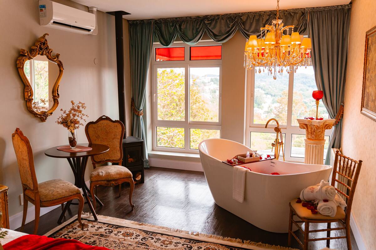 A room with a large bathtub, a chandelier, and a view of trees through a window.