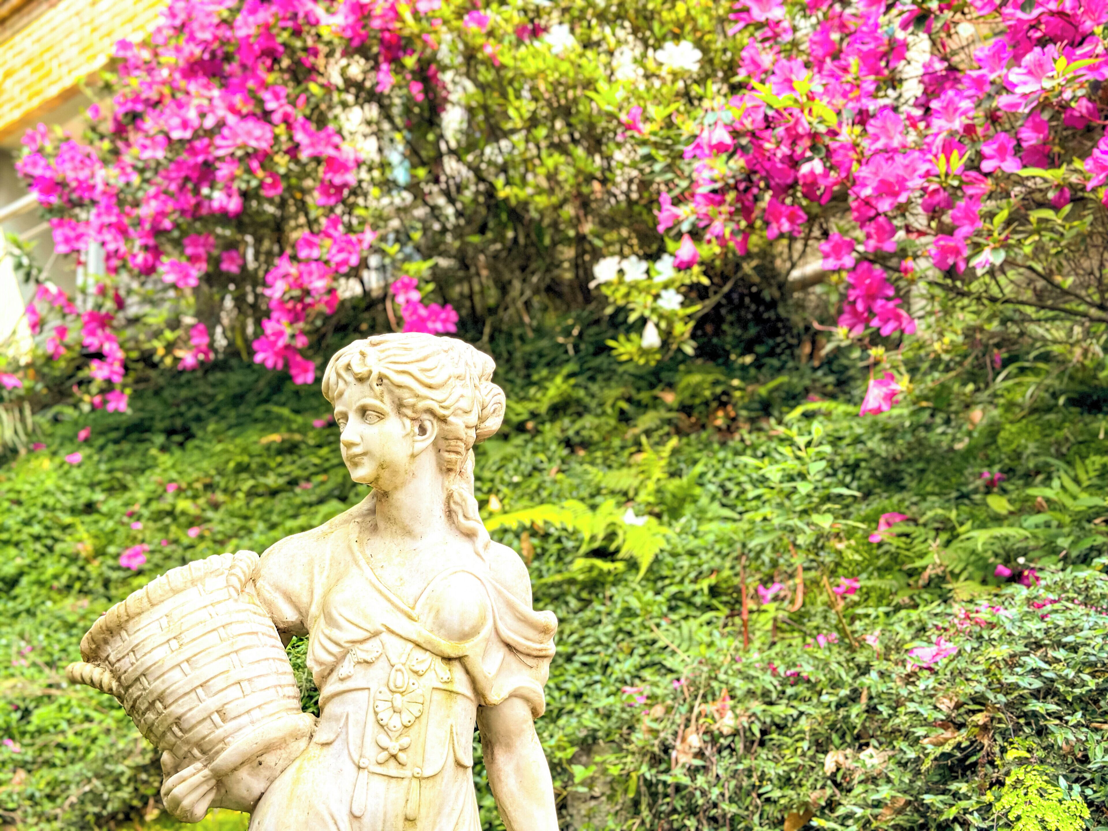 A statue of a woman holding a basket in a garden with pink flowers and greenery.