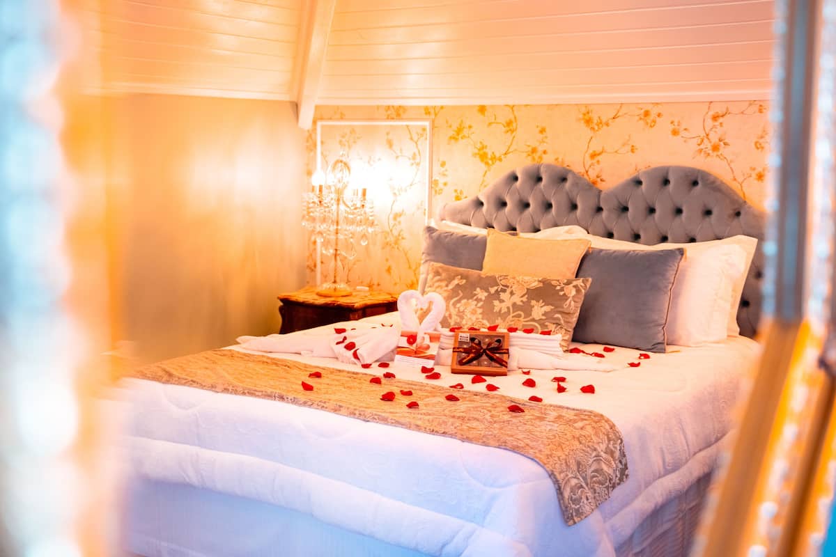 A neatly made bed with a grey tufted headboard, white linens, and rose petals scattered across the bed.