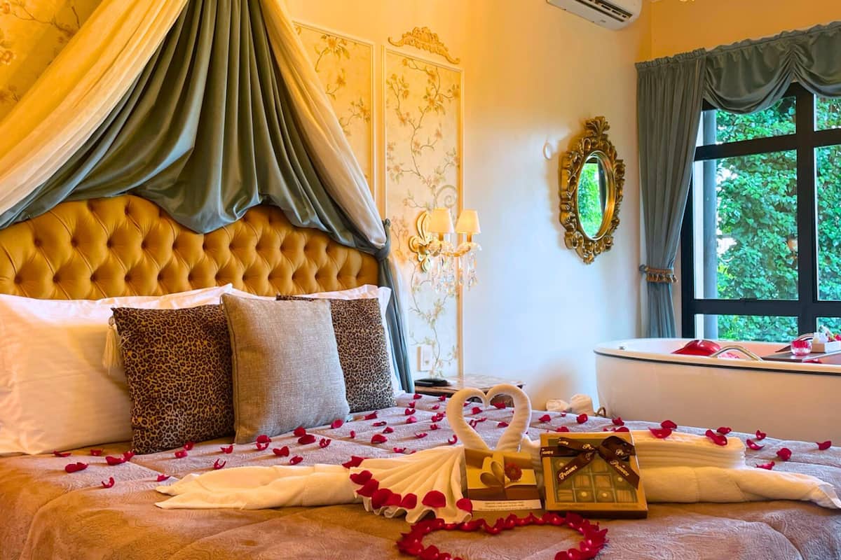 A luxurious bedroom with a tufted headboard, a large mirror, and a view of greenery outside.