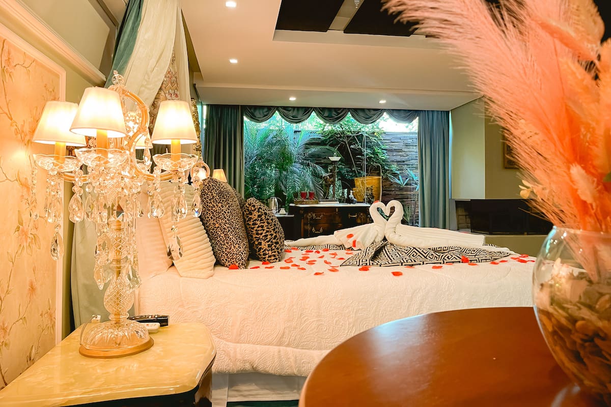A bedroom with a bed, a chandelier, a vase with flowers, and a view of greenery outside.