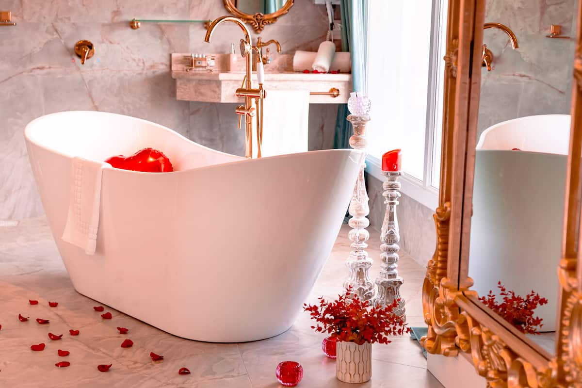 A white freestanding bathtub with a towel, a tall glass candle holder, and scattered red rose petals on a marble floor.
