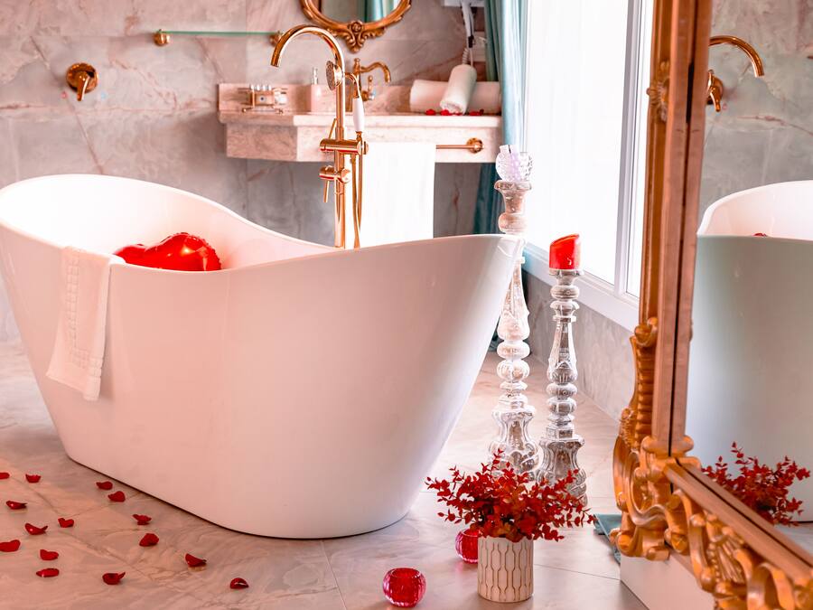 Une baignoire blanche, autoportante, avec une serviette, un grand porte-bougie en verre et des pétales de rose rouge éparpillés sur un sol en marbre.